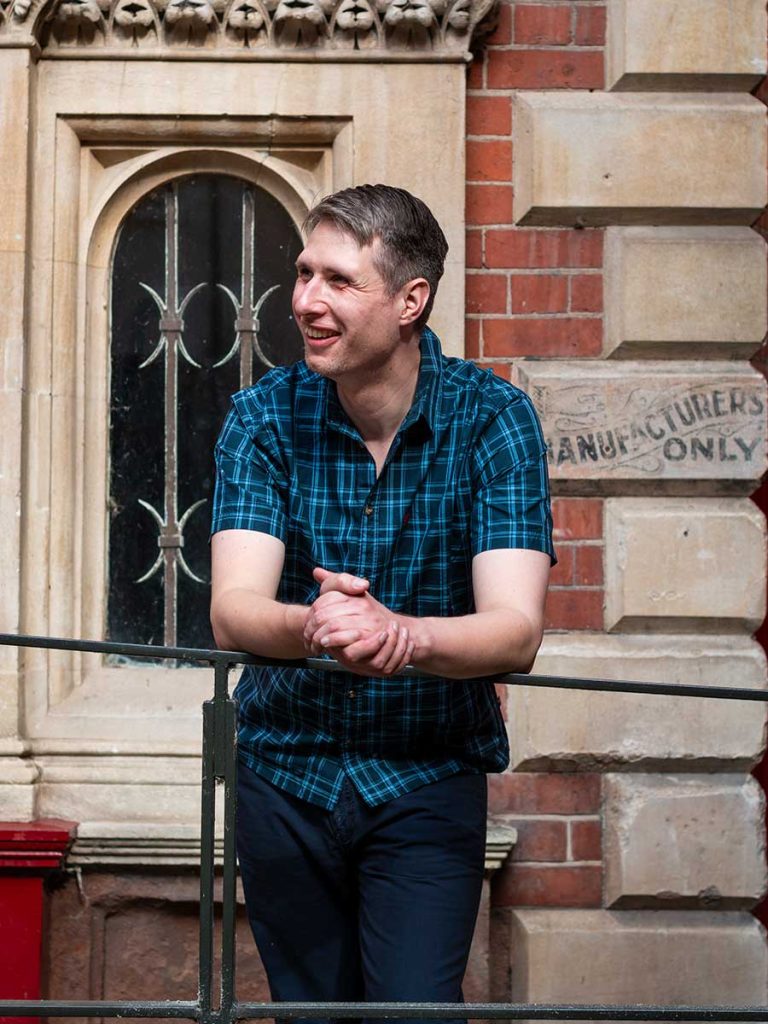 Dating Photo of male leaning on railing. He wears dark jeans and a navy and teal checked shirt.